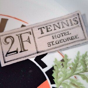 Op de afbeelding: Een wit stoffen label met zwarte tekst die "2F Tennis Hotel St. George" zegt.