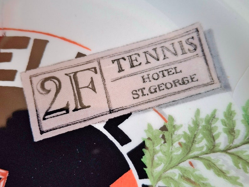 Op de afbeelding: Een wit stoffen label met zwarte tekst die "2F Tennis Hotel St. George" zegt.