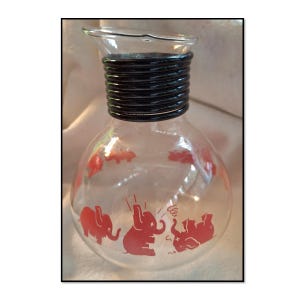 May include: A clear glass pitcher with a rounded body and a black, ridged neck. The pitcher features red elephant illustrations around the body. The spout is clear glass. The pitcher is likely vintage.