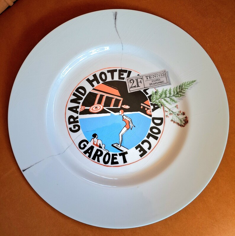 Op de afbeelding: Een wit keramiek bord met een blauw en rood ontwerp met de tekst "Grand Hotel La Dolce Garot" en een zwembadsc&egrave;ne. Een klein papieren kaartje met de tekst "Tennis Hotel St. George" en het nummer "2F" ligt op het bord.