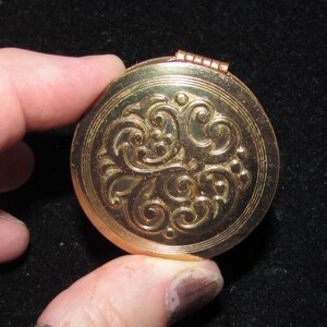 May include: A gold-colored compact with a decorative floral design. The compact has a hinged lid and a round shape.