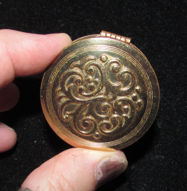 May include: A gold-colored compact with a decorative floral design. The compact has a hinged lid and a round shape.