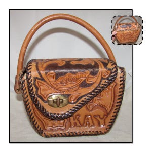 May include: A tan leather handbag with a curved handle and a decorative flap closure. The bag features embossed floral and geometric patterns, with the name "KAY" embossed on the front. The edges are trimmed with a dark brown braided design.