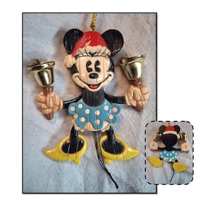 May include: A Minnie Mouse Christmas ornament. The ornament features Minnie Mouse wearing a Santa hat and a blue dress with white polka dots. She is holding a gold bell in each hand. Her shoes are yellow.