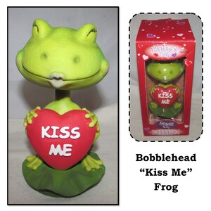 May include: A green frog bobblehead holding a red heart that says "Kiss Me". The frog is wearing a green lily pad base. The bobblehead is packaged in a box that says "Valentine's Day" and "Frances the Frog Bobblehead".