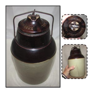 May include: Antique stoneware crock with a dark brown top and a lighter brown base. The lid has a metal handle and the text "THE WEIR" and "PAT. MAR 1ST 1892". The crock has a cylindrical shape and is likely used for storage.