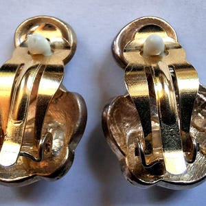 Puede incluir: Un par de pendientes vintage de clip. Los pendientes tienen un acabado met&aacute;lico dorado con un dise&ntilde;o floral. El mecanismo de clip es visible y los pendientes se muestran desde la parte posterior.