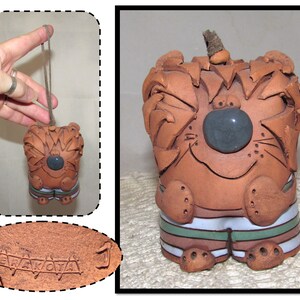 May include: A terracotta ceramic ornament of a cartoon animal with a brown, white, and green striped outfit. The ornament has a gray stone nose and a loop for hanging. The ornament is signed "TERRAKO'TA" on the bottom.