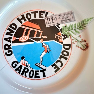 Op de afbeelding: Een wit keramiek bord met een kleurrijk ontwerp met een zwembad, twee figuren en de tekst "Grand Hotel Garot Dolce 2F Tennis Hotel St. George".