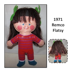 May include: A vintage 1971 Remco Flatsy doll. The doll has a painted face with blue eyes, pink cheeks, and a red smile. It wears a red and purple striped top and red pants, with blue shoes. Green hair accents are on the head.