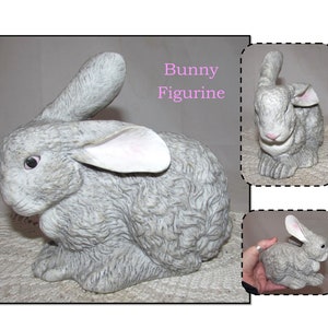 May include: A gray ceramic bunny figurine with pink ears. The figurine is sitting on a white surface. The text "Bunny Figurine" is written in pink letters above the figurine.