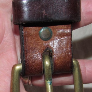 May include: A brown leather belt with a brass buckle. The belt has two decorative rivets with blue accents.