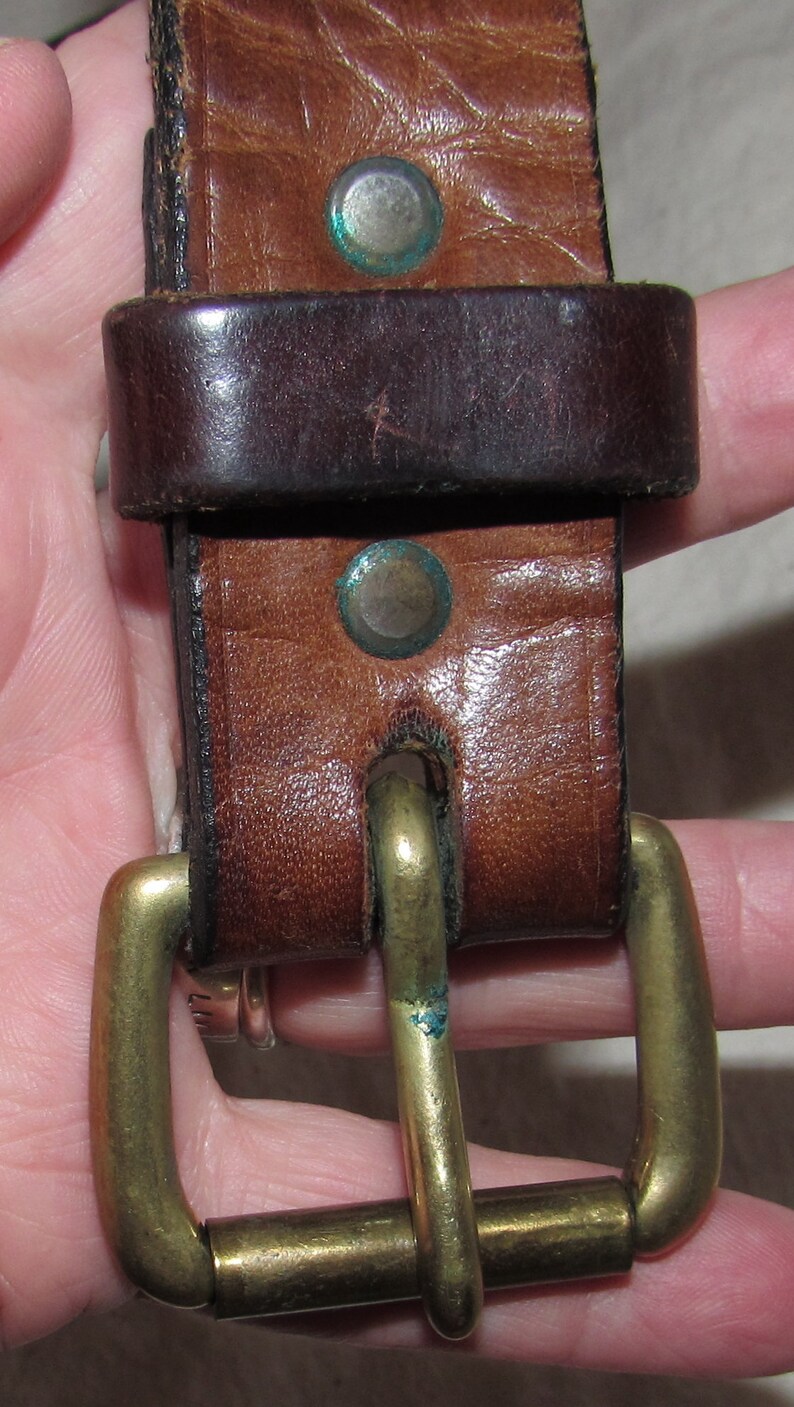 May include: A brown leather belt with a brass buckle. The belt has two decorative rivets with blue accents.