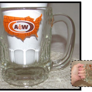 May include: A clear glass mug with a handle. The mug has a raised design around the bottom and a sticker on the front that says "A&W" with a map of the United States in the background.