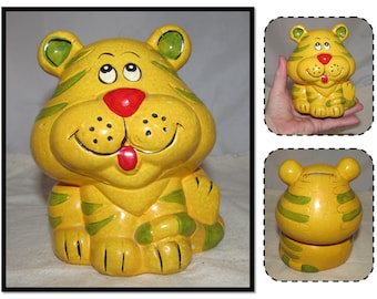 May include: A yellow ceramic piggy bank shaped like a cartoon tiger with green stripes. The tiger has a red nose and a big smile. The piggy bank has a slot on the back for coins.