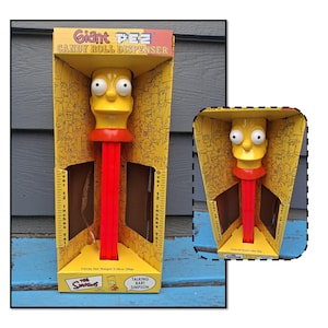 May include: A yellow and red Giant PEZ candy roll dispenser featuring Bart Simpson. The dispenser is in its original box, which is yellow with cartoon character illustrations. The box is over 12 inches tall.