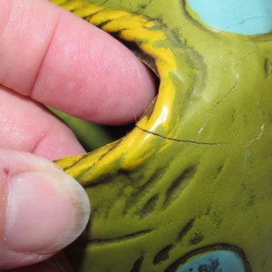 Pu&ograve; includere: Primo piano di un salvadanaio in ceramica verde, giallo e blu con una crepa sul lato. Il salvadanaio ha una grande bocca e un muso dipinto di giallo.