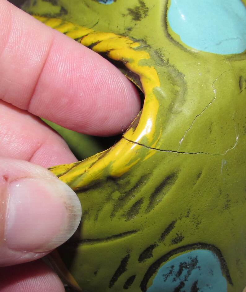 Pu&ograve; includere: Primo piano di un salvadanaio in ceramica verde, giallo e blu con una crepa sul lato. Il salvadanaio ha una grande bocca e un muso dipinto di giallo.