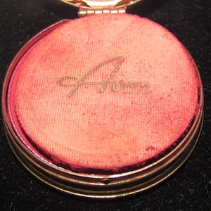 May include: A gold-colored compact with a red velvet interior. The interior is inscribed with the word "Avon" in gold lettering.