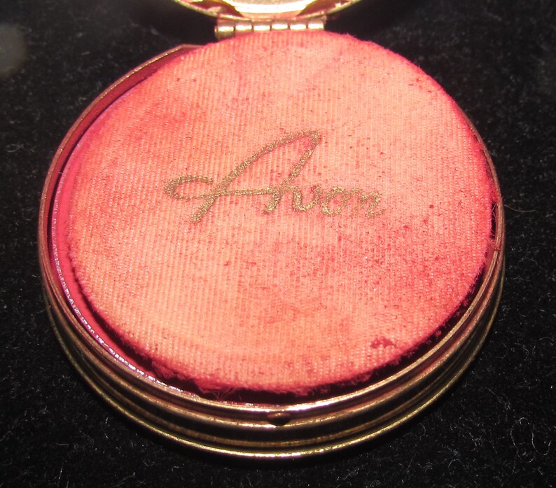 May include: A gold-colored compact with a red velvet interior. The interior is inscribed with the word "Avon" in gold lettering.