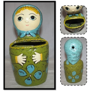 Pu&ograve; includere: Un vaso da fiori in ceramica a forma di donna con un foulard blu e un vestito verde. Il vaso ha una fessura nella parte anteriore per contenere piccoli oggetti.