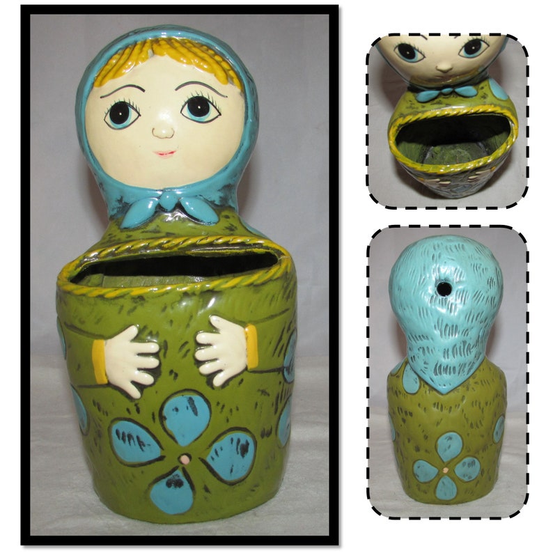 Pu&ograve; includere: Un vaso da fiori in ceramica a forma di donna con un foulard blu e un vestito verde. Il vaso ha una fessura nella parte anteriore per contenere piccoli oggetti.