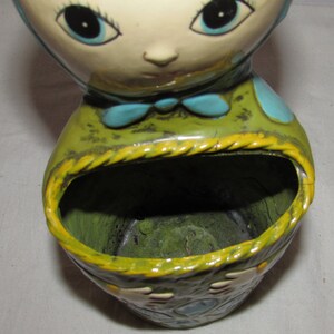 Pu&ograve; includere: Un vaso da fiori in ceramica a forma di testa di bambola con grandi occhi blu e un corpo con motivi verdi e blu. Il vaso ha un'ampia apertura nella parte superiore per la semina.