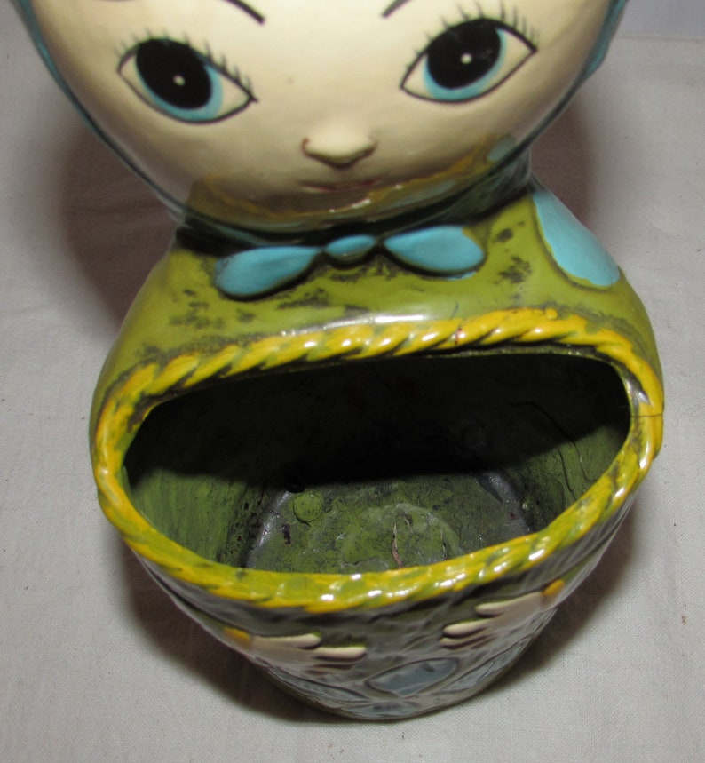 Pu&ograve; includere: Un vaso da fiori in ceramica a forma di testa di bambola con grandi occhi blu e un corpo con motivi verdi e blu. Il vaso ha un'ampia apertura nella parte superiore per la semina.