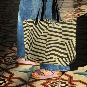 May include: A large tote bag with a black and tan striped pattern is held by a person wearing blue jeans and pink sandals. The bag has black leather handles and is made of a sturdy fabric.