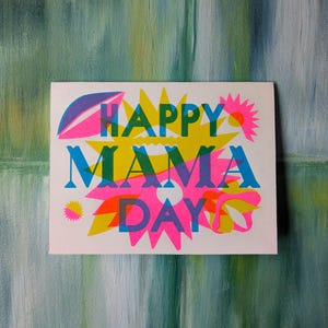 May include: A white card with the words "HAPPY MAMA DAY" in overlapping neon colors. The design includes yellow starbursts, pink flowers, and blue and green accents. The background is a textured, abstract painting.