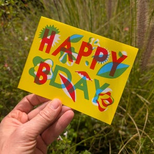 May include: A yellow birthday card with the words "HAPPY BDAY" in red, green, and blue. The card has a floral design with a green and blue leaf pattern. The card is held in a hand.