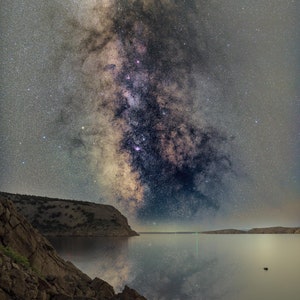 May include: A night sky with the Milky Way galaxy visible above a body of water. The galaxy is a bright band of stars and gas, stretching across the sky. The water is calm and reflects the stars.