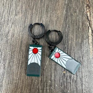 Può includere: Un paio di orecchini a cerchio neri con pendenti rettangolari. Ogni pendente presenta un disegno di sole rosso con raggi bianchi su sfondo verde scuro. Gli orecchini sono esposti su una superficie di legno.