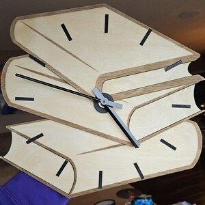 May include: A wooden clock shaped like a stack of books with black hour and minute hands. The clock face is made of light wood with black markings for the hours.