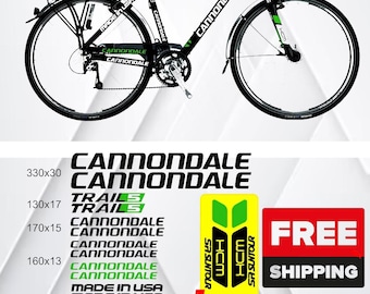 Könnte beinhalten: Schwarzes Cannondale Trails Fahrrad mit grünen Akzenten. Es verfügt über einen Gepäckträger, Schutzbleche und die Schriftzüge "Cannondale" und "Trails". Enthält Aufkleber für die Gabel und den Text "Made in USA".