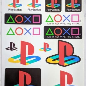 May include: A sheet of stickers featuring various PlayStation logos, including the iconic blue, red, yellow, and green PlayStation logo, the PlayStation text logo, and the PlayStation button symbols: triangle, circle, cross, and square. The sheet also includes the slogan "Live In Your World, Play In ours."