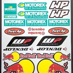 Peut inclure: Un ensemble d'autocollants de motocross avec différentes marques et logos, notamment Acerbis, Renthal, D.I.D., WP, Motorex, Twin Air, Brembo et Red Bull. Les autocollants sont imprimés sur un fond blanc.