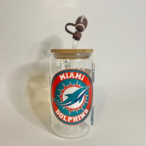 May include: A clear glass water bottle with a bamboo lid and a silicone straw with a football-shaped top. The bottle has a Miami Dolphins logo with the words "Miami Dolphins" printed on it.