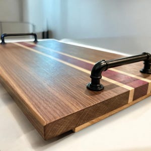Puede incluir: Bandeja rectangular de madera de nogal oscuro. Cuenta con una franja central de madera morada y crema, y asas de metal negro. La bandeja está diseñada para servir alimentos y bebidas.
