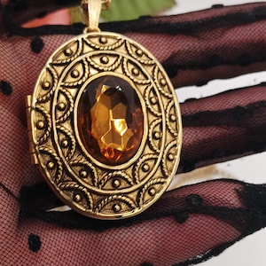 Op de afbeelding: Een ovaal medaillon-hangertje van goudkleurig metaal aan een bijpassende ketting. Het medaillon is voorzien van een ingewikkeld reliëfpatroon van concentrische cirkels en stippen rondom een grote, gefacetteerde, barnsteenkleurige ovale edelsteen.