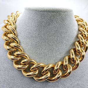 May include: A chunky gold-tone chain necklace. The necklace features large, interlocking links with a polished finish. The jewelry is displayed on a gray mannequin bust. The chain is a bold statement piece.