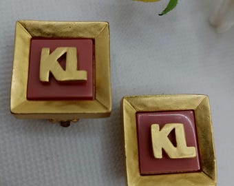 May include: A pair of square clip-on earrings. Each earring features a gold-colored frame surrounding a mauve-colored square. The letters "KL" are in gold within the mauve square. The earrings are on a white surface.
