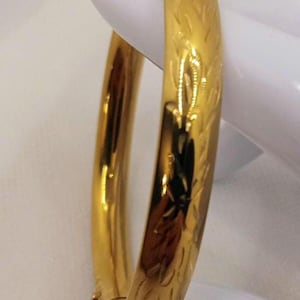 Könnte beinhalten: Ein polierter, goldfarbener Scharnierarmreif mit einem dezenten, sich wiederholenden, blattartigen Gravurmuster auf der Außenseite. Der Armreif hat ein glattes, abgerundetes Profil und einen sicheren Verschlussmechanismus am unteren Band.
