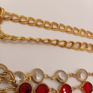 May include: Close-up of a gold-toned necklace with three strands. The top two strands are chain links, and the bottom strand features alternating red and clear gemstones set in gold. The necklace is on a white background.