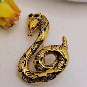 May include: A gold-toned metal brooch shaped like a coiled snake. The snake features a polished, reflective surface with recessed black decorative patterns along its body and head. It has two small, dark, circular eyes and a curved tail.