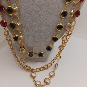 May include: Three gold-toned necklaces with round, bezel-set gemstones. The gemstones are clear, black, and red. The chains have a link design. The necklaces are displayed on a gray mannequin.