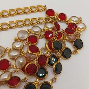 May include: Gold-tone necklace with a chain design and multiple strands of colorful, round gemstones. The gemstones are red, clear, and black. The necklace is on a white background.