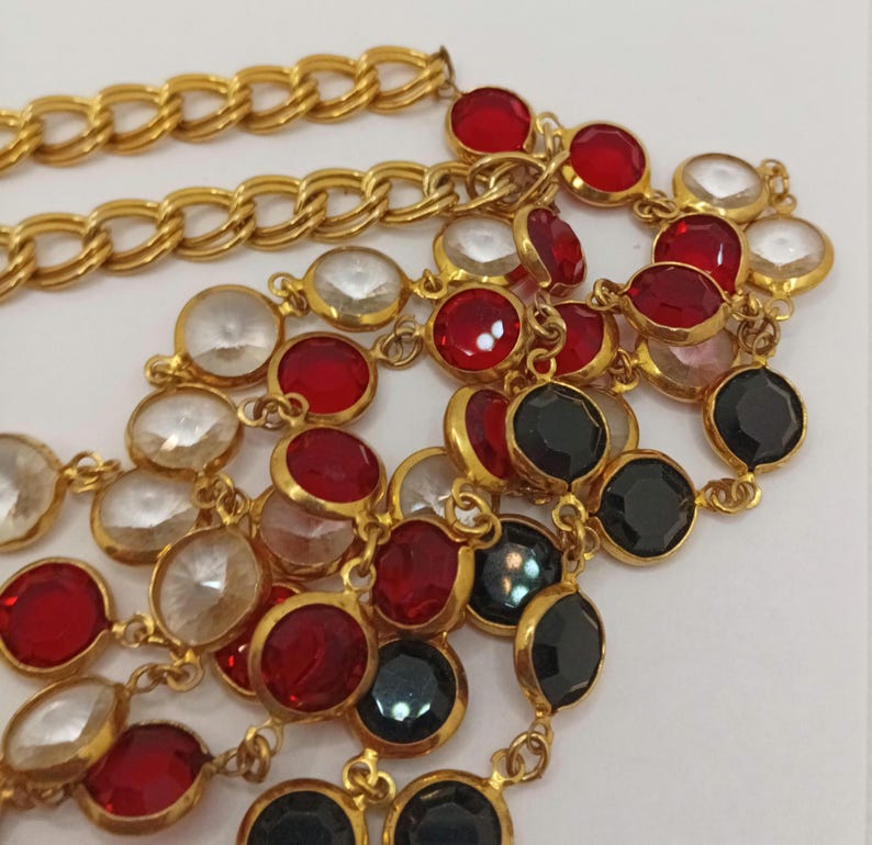 May include: Gold-tone necklace with a chain design and multiple strands of colorful, round gemstones. The gemstones are red, clear, and black. The necklace is on a white background.