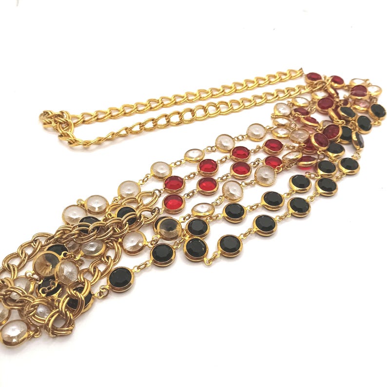 May include: A gold-toned necklace with multiple strands. Each strand features a series of round, colorful gemstones in clear, red, and black. The necklace has a chain closure.