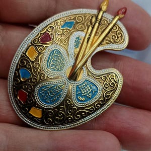 Puede incluir: Broche de paleta de artista detallado con detalles dorados y plateados. La paleta presenta manchas de pintura coloridas en rojo, amarillo y azul, con tres pinceles dorados. El diseño incluye intrincados adornos y un borde decorativo.